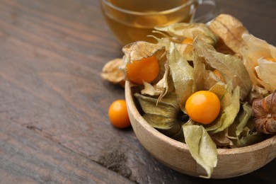 Ripe physalis fruits with calyxes in bowl on wooden table, closeup. Space for text