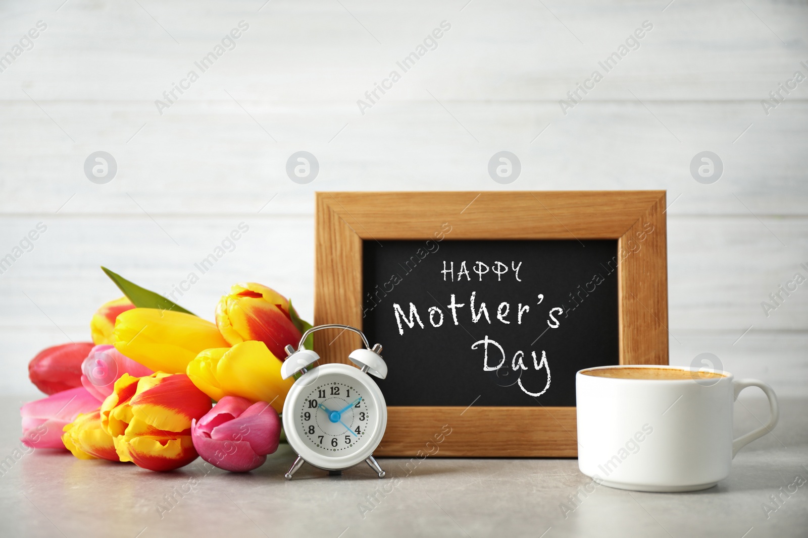 Image of Chalkboard with phrase HAPPY MOTHER'S DAY, tulips, cup of coffee and alarm clock on light table