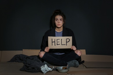 Photo of Poor young woman with HELP sign sitting on floor near dark wall