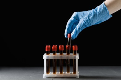 Scientist putting test tube with brown liquid into stand at grey table against black background, closeup