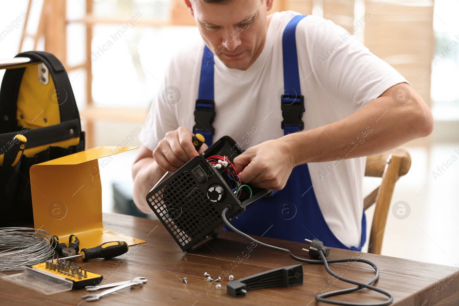 Photo of Professional technician repairing electric fan heater with screwdriver at table indoors