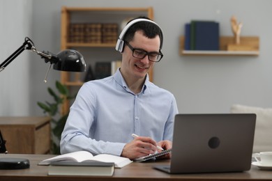 Photo of E-learning. Young man taking notes during online lesson at table indoors.