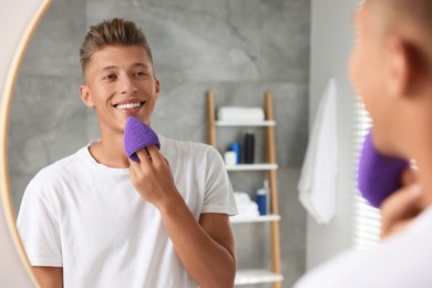 Happy young man washing his face with sponge near mirror in bathroom