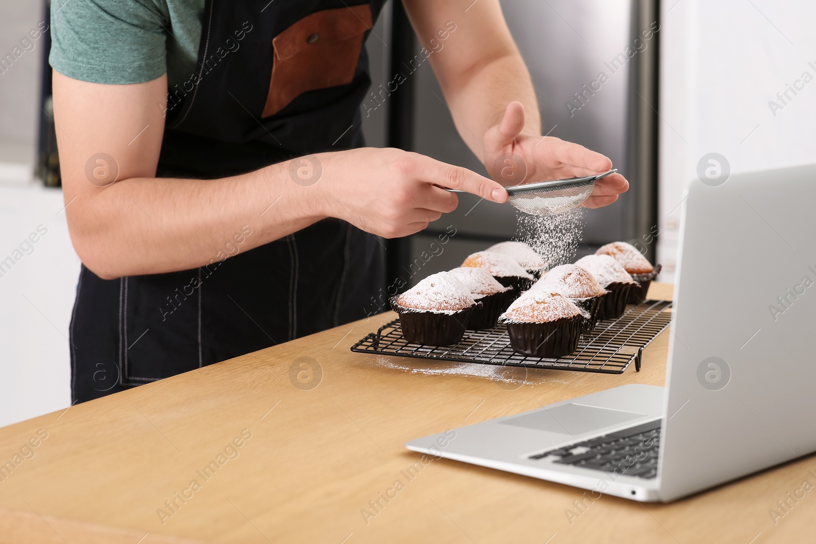 Photo of Man decorating muffins with powdered sugar while watching online cooking course via laptop in kitchen, closeup. Time for hobby