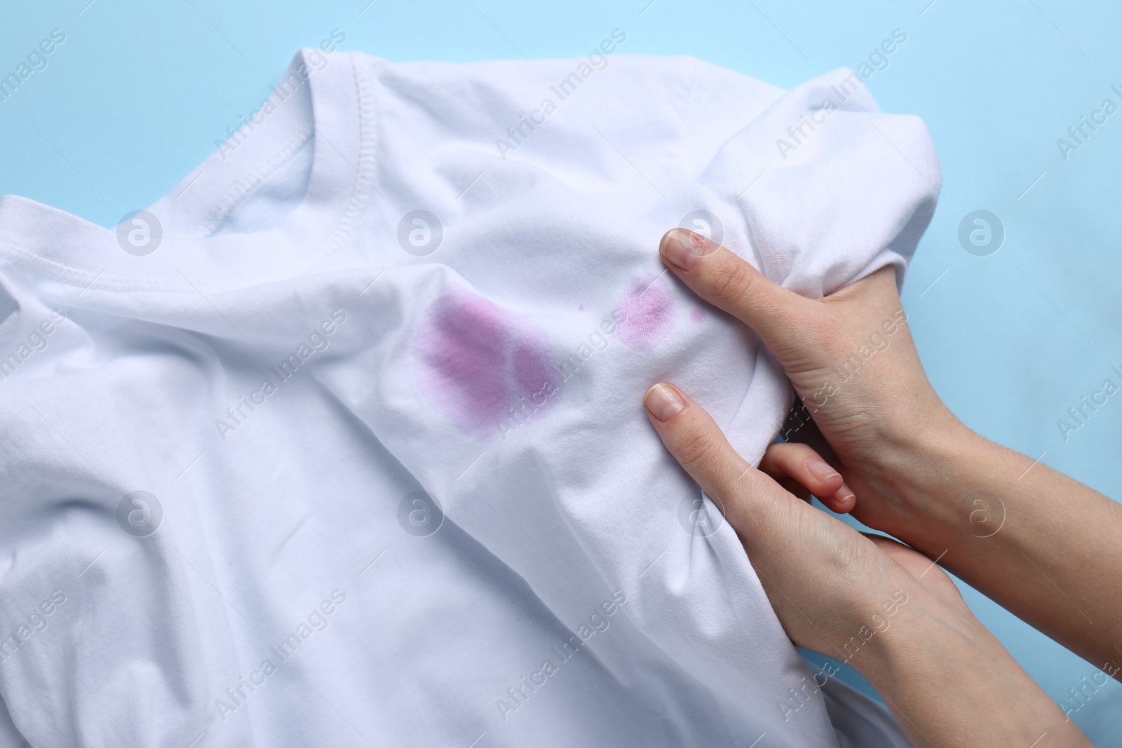 Photo of Woman holding white shirt with purple stains on light blue background, top view