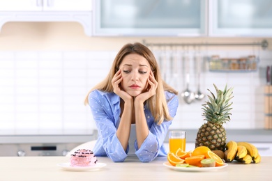 Woman choosing between dessert and fruits at table in kitchen. Healthy diet