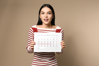Young woman holding calendar with marked menstrual cycle days on beige background