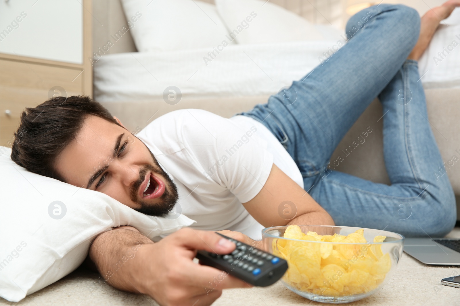 Photo of Lazy young man with bowl of chips watching TV while lying on floor at home