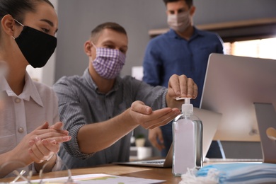 Photo of Coworkers with protective masks using hand sanitizer in office. Business meeting during COVID-19 pandemic