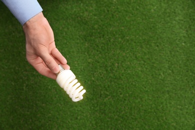 Man holding energy saving bulb for lamp over green grass, top view. Space for text
