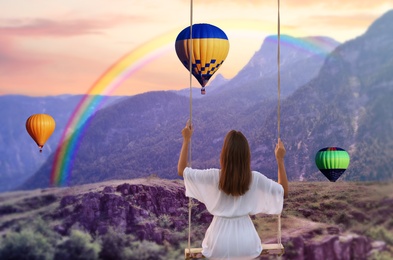 Dream world. Young woman swinging over mountains, hot air balloons in sunset sky on background