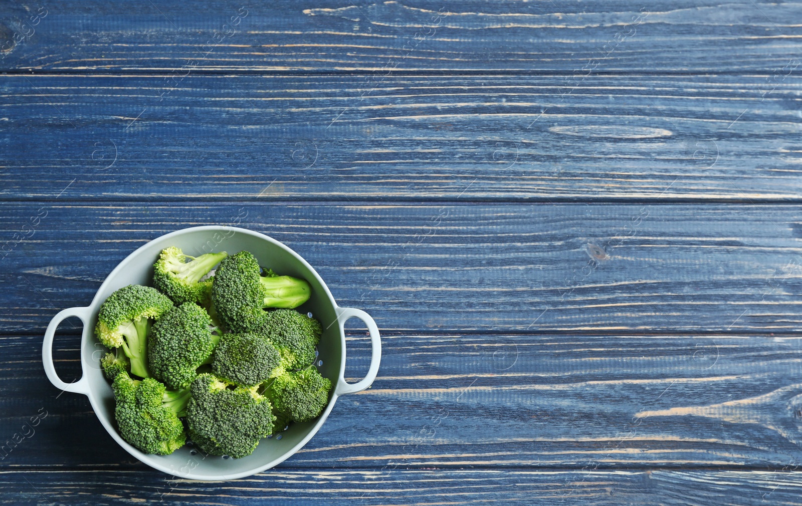 Photo of Colander of fresh broccoli on blue wooden table, top view with space for text