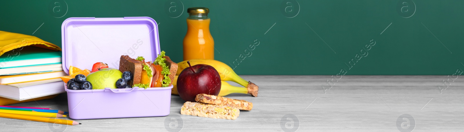 Image of School meal. Lunch box with healthy food and different stationery on light wooden table near green chalkboard, space for text. Banner design