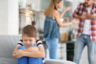 Little unhappy boy sitting on sofa while parents arguing at home