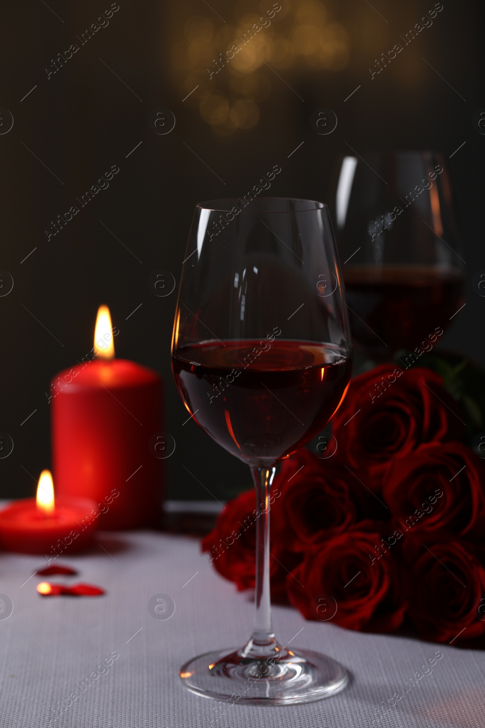 Photo of Glasses of red wine, burning candles and rose flowers on grey table against blurred lights. Romantic atmosphere