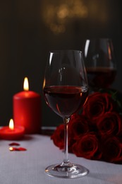 Glasses of red wine, burning candles and rose flowers on grey table against blurred lights. Romantic atmosphere