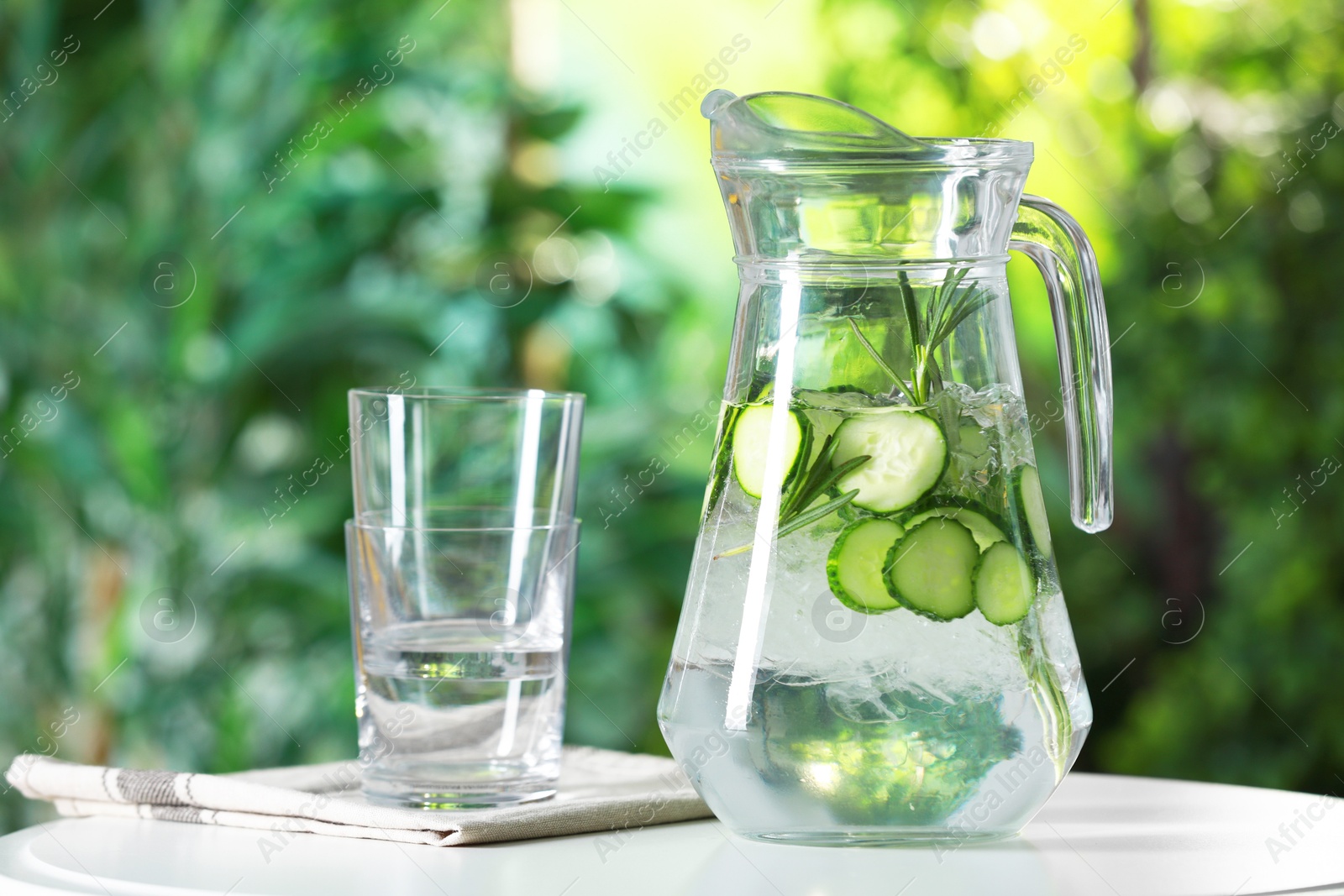 Photo of Refreshing cucumber water with rosemary in jug and glasses on white table against blurred green background, closeup