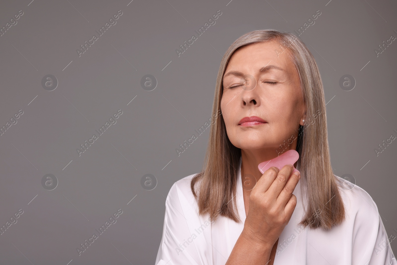 Photo of Woman massaging her face with rose quartz gua sha tool on grey background, space for text