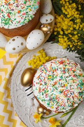 Traditional Easter cakes with sprinkles, painted eggs and beautiful spring flowers on yellow background, flat lay