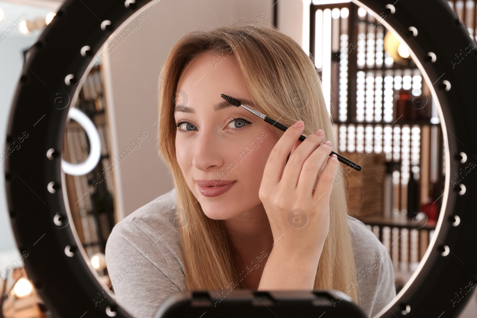 Photo of Beautiful young woman brushing eyebrows indoors, view through ring lamp