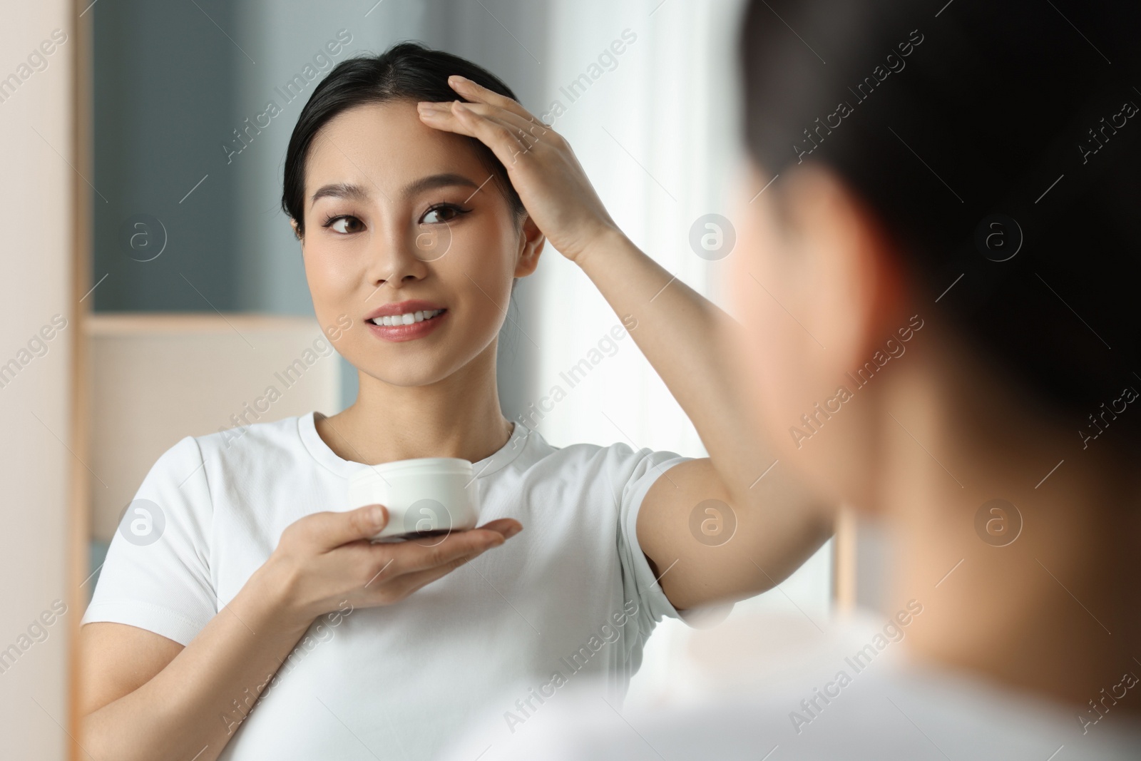 Photo of Happy woman applying face cream near mirror at home