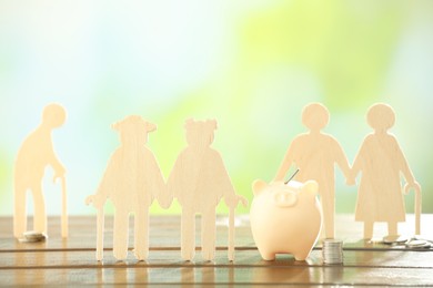 Photo of Pension savings. Figures of elderly people, piggy bank and coins on wooden table against blurred background