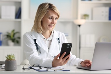 Photo of Smiling doctor with smartphone and laptop having online consultation at table in office