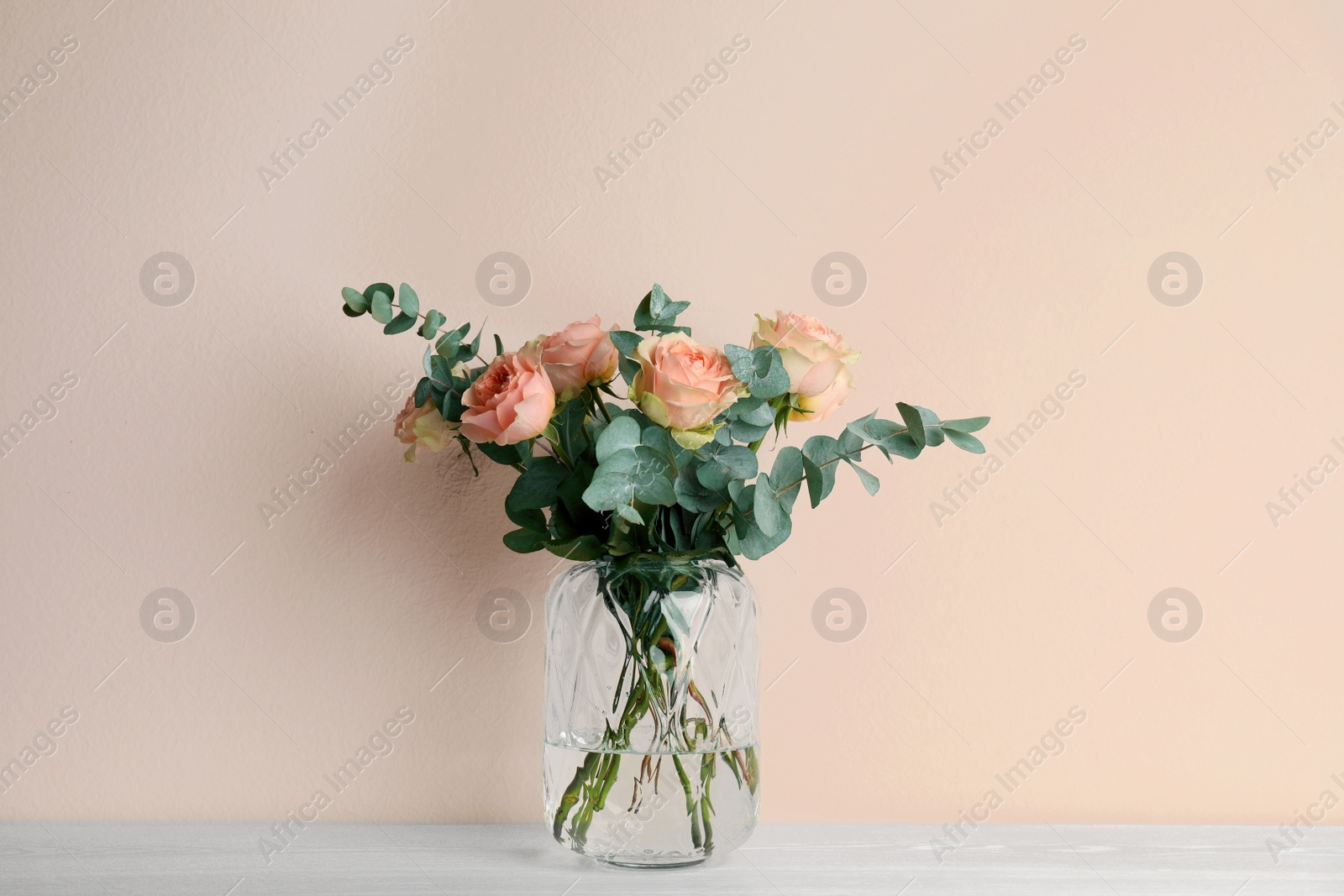 Photo of Vase with bouquet of beautiful roses on white wooden table near beige wall