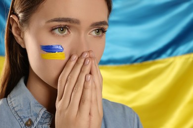 Photo of Sad young woman with clasped hands near Ukrainian flag, closeup. Space for text