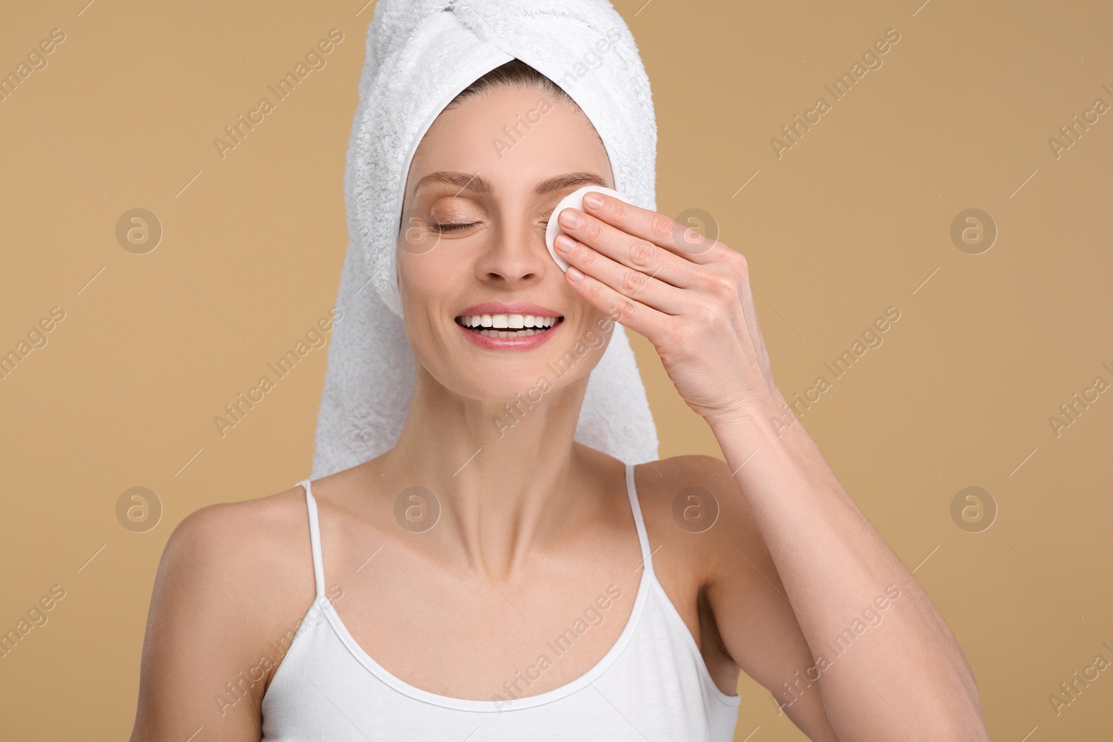 Photo of Beautiful woman in terry towel removing makeup with cotton pad on beige background