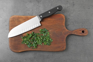 Wooden board with sharp knife and chopped parsley on grey background, top view