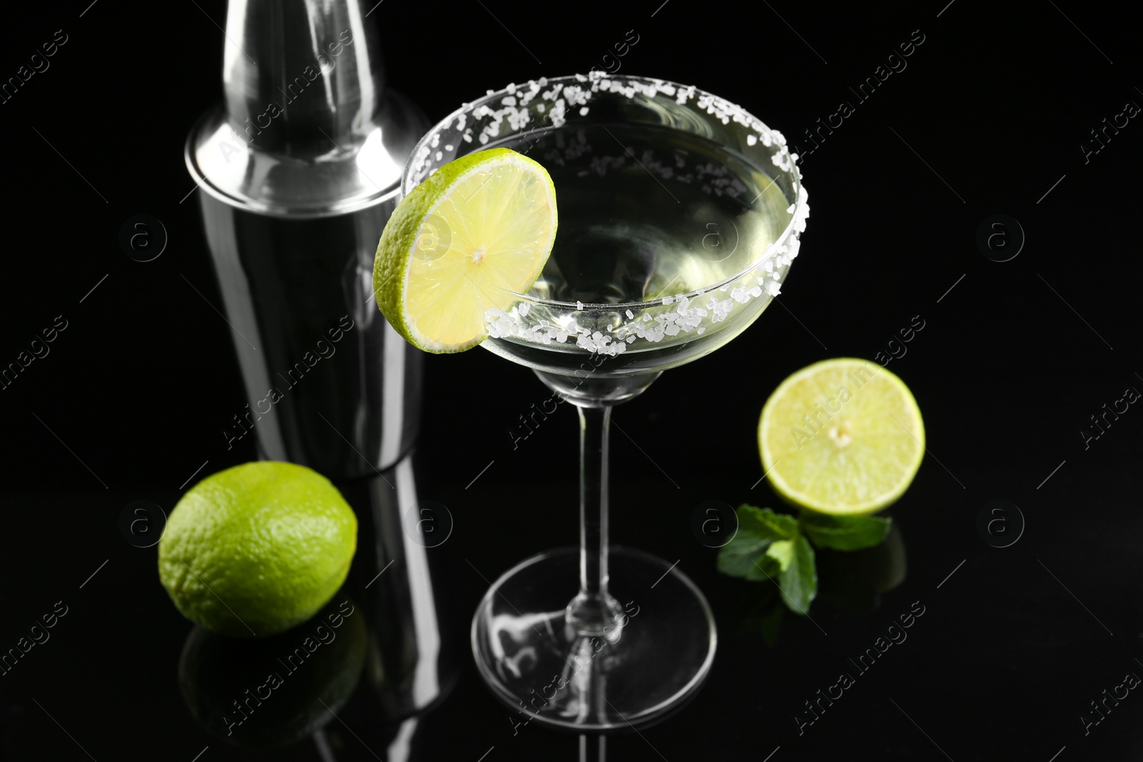 Photo of Metal shaker, delicious cocktail, limes and mint on black mirror surface