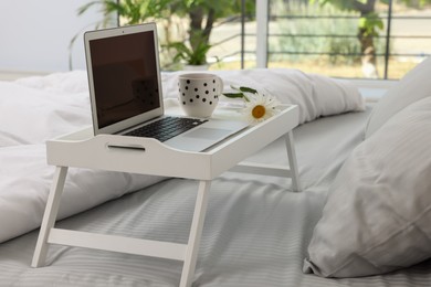Photo of White tray table with laptop, cup of drink and daisy on bed indoors