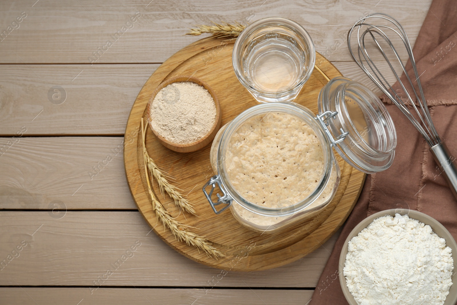 Photo of Leaven, ears of wheat, whisk, water and flour on beige wooden table, flat lay