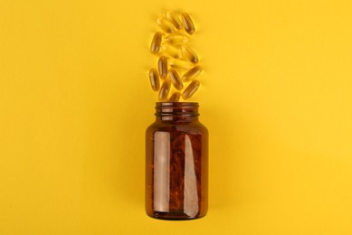 Photo of Glass medical bottle and vitamin capsules on yellow background, top view