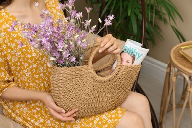 Woman holding beach bag with beautiful bouquet of wildflowers and magazine indoors, closeup