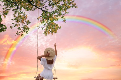 Image of Dream world. Young woman swinging, rainbow in sunset sky on background