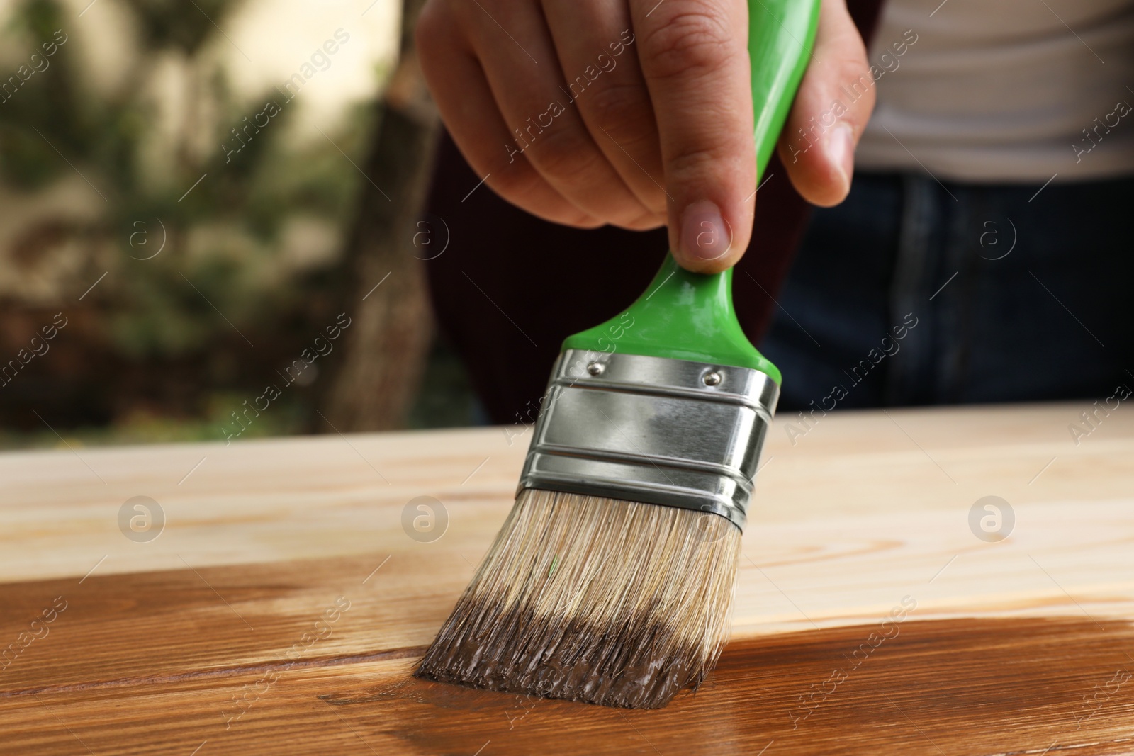 Photo of Man applying wood stain onto wooden surface against blurred background, closeup