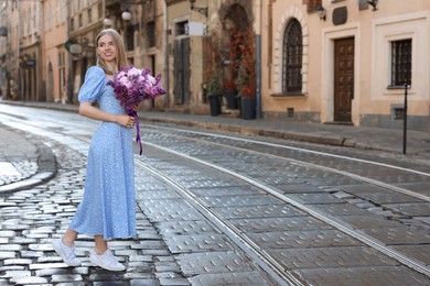 Beautiful woman with bouquet of spring flowers on city street, space for text