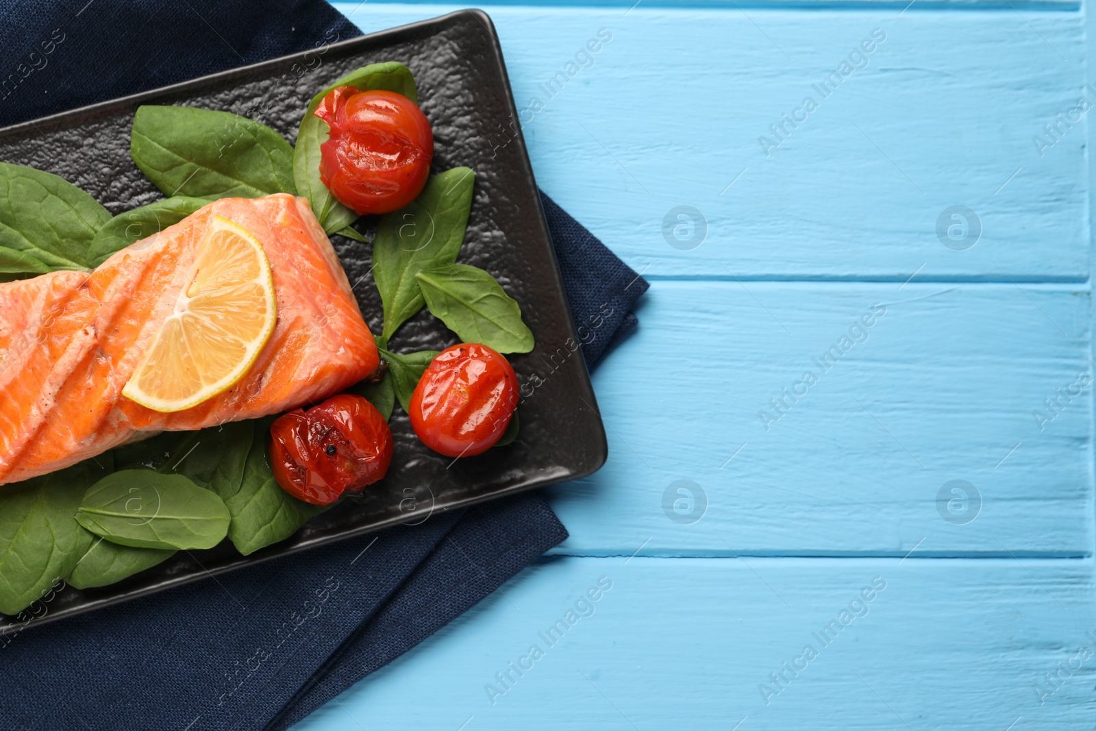 Photo of Tasty grilled salmon with tomatoes, spinach and lemon on light blue wooden table, top view. Space for text