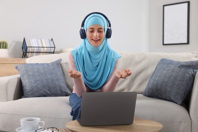 Photo of Muslim woman in headphones using video chat on laptop at wooden table in room
