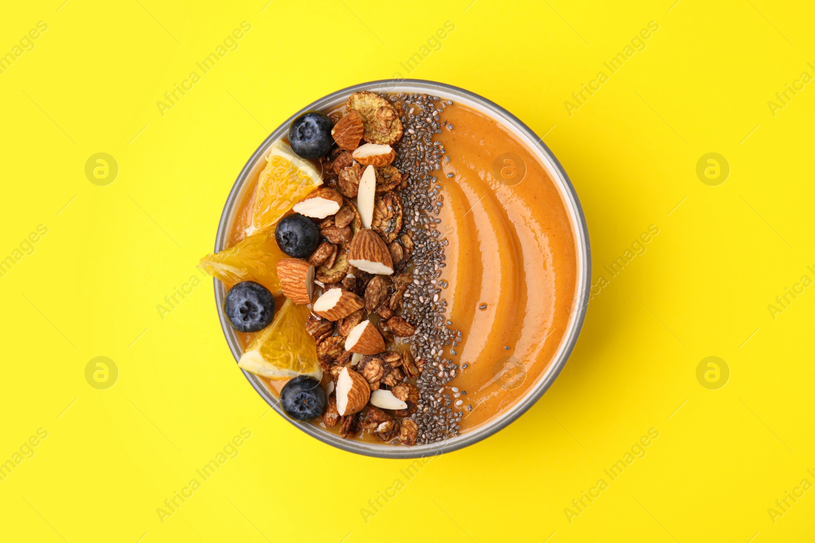 Photo of Bowl of delicious fruit smoothie with fresh orange slices, blueberries and granola on yellow background, top view