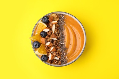 Bowl of delicious fruit smoothie with fresh orange slices, blueberries and granola on yellow background, top view