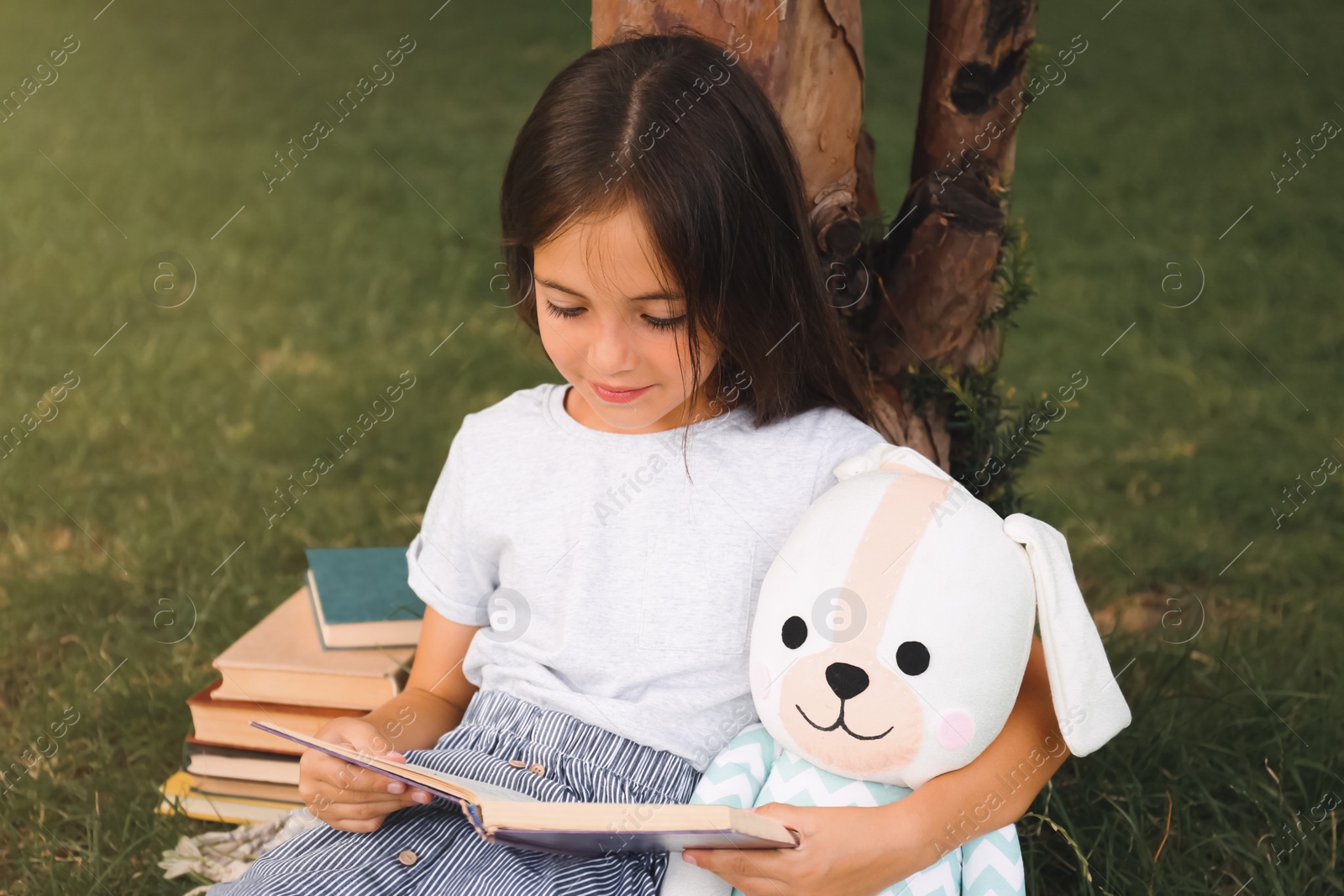 Photo of Cute little girl with toy reading book on green grass near tree in park