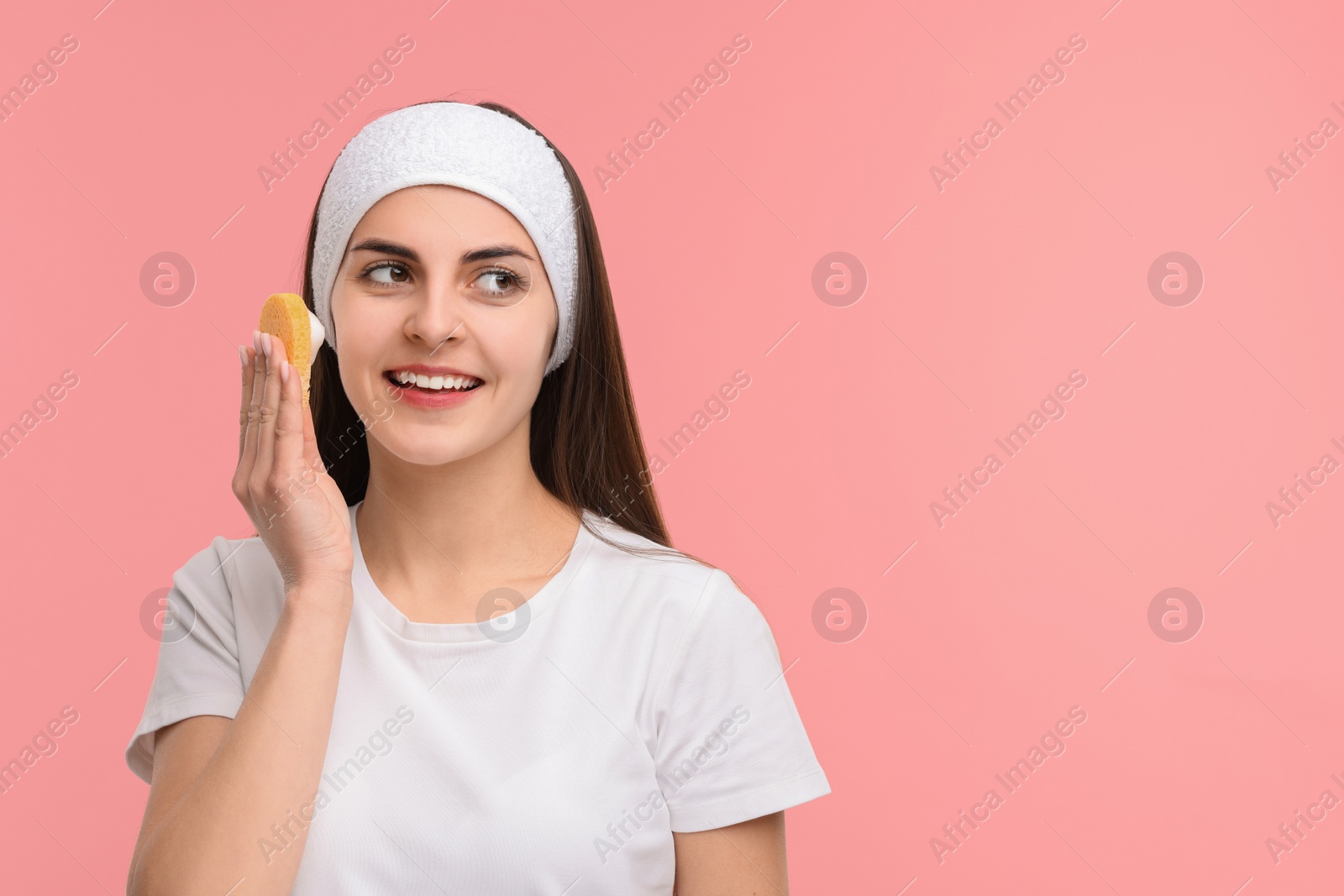 Photo of Young woman with headband washing her face using sponge on pink background, space for text