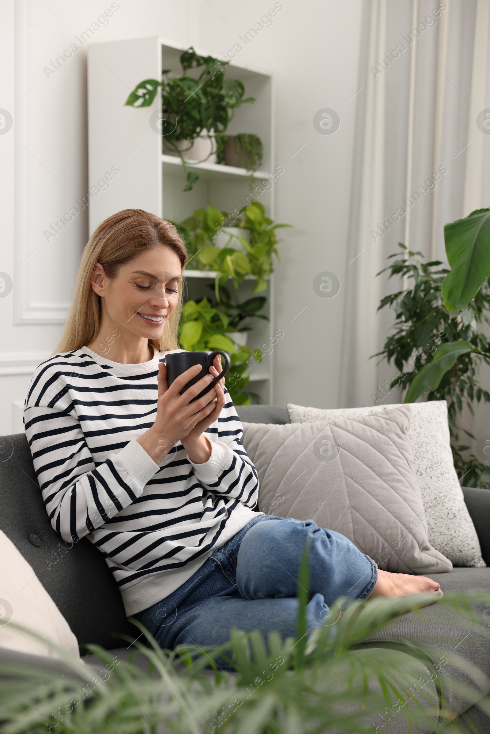 Photo of Woman with cup of drink sitting on sofa surrounded by beautiful potted houseplants at home