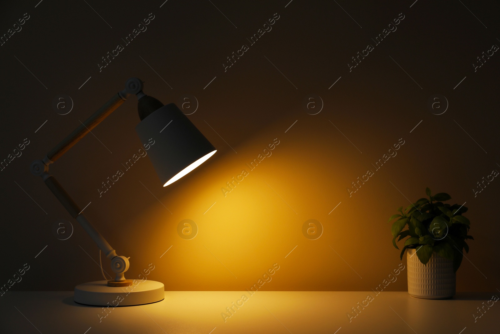 Photo of Stylish modern desk lamp and potted plant on table near wall in dark room