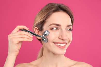 Photo of Young woman using metal face roller on pink background