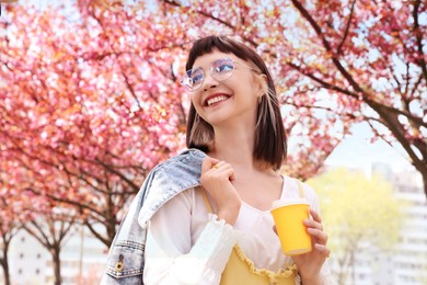 Beautiful young woman holding paper coffee cup in park with blossoming sakura trees