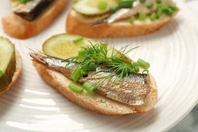 Photo of Delicious sandwiches with sprats, pickled cucumber, green onion and dill on plate, closeup